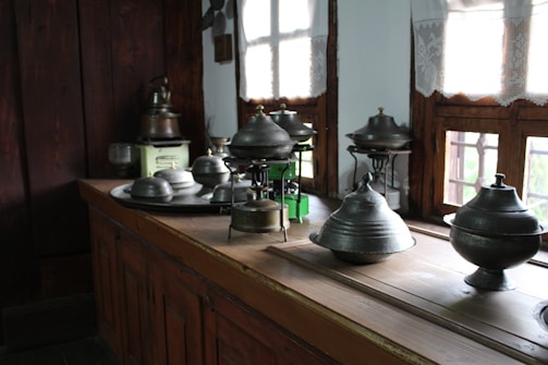 A collection of metal kitchenware, including various pots and lids, is arranged on a wooden kitchen counter. The room features wooden walls and windows with light curtains, allowing natural sunlight to illuminate the space.