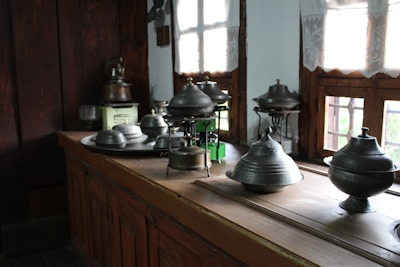 A collection of metal kitchenware, including various pots and lids, is arranged on a wooden kitchen counter. The room features wooden walls and windows with light curtains, allowing natural sunlight to illuminate the space.