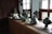A collection of metal kitchenware, including various pots and lids, is arranged on a wooden kitchen counter. The room features wooden walls and windows with light curtains, allowing natural sunlight to illuminate the space.