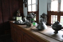 A collection of metal kitchenware, including various pots and lids, is arranged on a wooden kitchen counter. The room features wooden walls and windows with light curtains, allowing natural sunlight to illuminate the space.