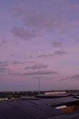 A rooftop covered with solar panels stretching towards the horizon under a pastel pink and purple sky. Fluffy clouds drift across the sky with a small moon visible amidst them. The horizon features a distant cityscape with a subtle blend of green trees and urban structures.