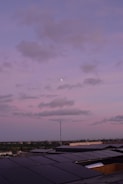 A rooftop covered with solar panels stretching towards the horizon under a pastel pink and purple sky. Fluffy clouds drift across the sky with a small moon visible amidst them. The horizon features a distant cityscape with a subtle blend of green trees and urban structures.