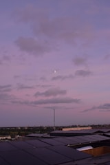 A rooftop covered with solar panels stretching towards the horizon under a pastel pink and purple sky. Fluffy clouds drift across the sky with a small moon visible amidst them. The horizon features a distant cityscape with a subtle blend of green trees and urban structures.