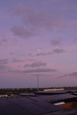 A rooftop covered with solar panels stretching towards the horizon under a pastel pink and purple sky. Fluffy clouds drift across the sky with a small moon visible amidst them. The horizon features a distant cityscape with a subtle blend of green trees and urban structures.