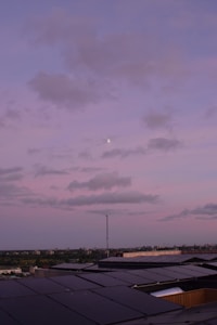 A rooftop covered with solar panels stretching towards the horizon under a pastel pink and purple sky. Fluffy clouds drift across the sky with a small moon visible amidst them. The horizon features a distant cityscape with a subtle blend of green trees and urban structures.