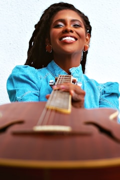 A person smiling widely while holding a guitar. The focus is on the joyful expression and the blue denim shirt worn by the person. The image captures a sense of happiness and warmth.