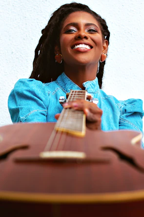 A student holding a guitar, smiling, with a campus building blurred in the background