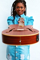 An individual with long braided hair smiles while holding a guitar towards the camera, dressed in a blue denim outfit with torn patches.