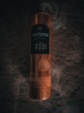 A cylindrical container with a tartan pattern, featuring striking orange hues and the label '100 Pipers Exceptional Blended Scotch Whisky'. The packaging includes embossed details and a seal of Scotland indicating its origin.