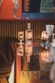A bar setting featuring a bottle of Anae liquor and a cocktail glass with a garnish, placed on a polished wooden bar counter. The background includes colorful vintage posters and a warm ambient light adds a cozy atmosphere.