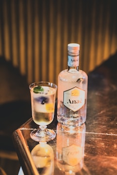 A bottle of Anaë gin and a cocktail glass are placed on a reflective surface. The glass contains a floral garnish along with a slice of lemon and some ice cubes. The background has a warm, dimly lit setting, creating a cozy ambiance.