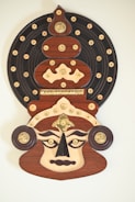 A highly stylized and decorative mask that features intricate patterns of gold embellishments on a dark, circular base. The mask itself has exaggerated facial features with large, almond-shaped eyes, a prominent pointed nose, and a frowning expression. The color palette includes dark brown, gold, and ivory, creating a striking contrast.