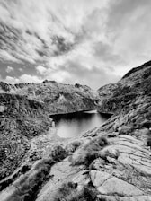 A black and white landscape featuring a mountainous region with rugged terrain and a tranquil lake nestled between rocky peaks. The sky is partly cloudy, adding texture and depth to the scene. The foreground includes patches of grass and rocky outcrops, leading the eye towards the serene water.
