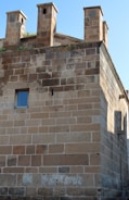 A stone building with three rectangular chimneys on the roof. The wall is constructed from large, unevenly colored stone blocks. A single small window with reflective glass is set into the wall. Vegetation grows at the top.