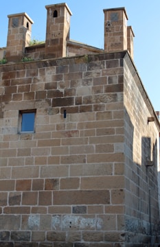 A stone building with three rectangular chimneys on the roof. The wall is constructed from large, unevenly colored stone blocks. A single small window with reflective glass is set into the wall. Vegetation grows at the top.