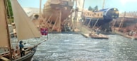 A detailed model depicting a busy shipyard with several ships in various stages of construction. The scene includes a small sailboat with a crew member holding a flag, rippling water, and workers on a wooden platform and onshore, surrounded by scaffolding and shipbuilding materials.