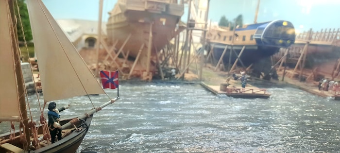 A detailed model depicting a busy shipyard with several ships in various stages of construction. The scene includes a small sailboat with a crew member holding a flag, rippling water, and workers on a wooden platform and onshore, surrounded by scaffolding and shipbuilding materials.