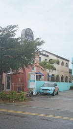 A pastel-colored building features a sign for Archie's of Siesta Key and appears to house a massage therapy business. A light blue Mini Cooper is parked in front of the building. The scene includes palm trees and a wet road, suggesting recent rain.