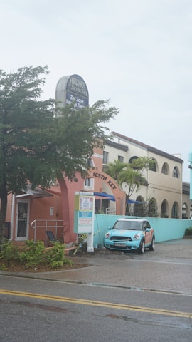 A pastel-colored building features a sign for Archie's of Siesta Key and appears to house a massage therapy business. A light blue Mini Cooper is parked in front of the building. The scene includes palm trees and a wet road, suggesting recent rain.