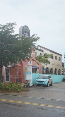 A pastel-colored building features a sign for Archie's of Siesta Key and appears to house a massage therapy business. A light blue Mini Cooper is parked in front of the building. The scene includes palm trees and a wet road, suggesting recent rain.