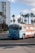 A vintage bus painted with a faded American flag design is parked in an open area. The bus is light blue with stars and red and white stripes. In the background, there are tall palm trees and a tall, modern apartment building. A small seagull is also visible to the side of the bus.