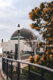 A large observatory building with a dome roof is seen in the background, surrounded by trees. The foreground features a railing and a lamp post, with some individuals walking and gathering outside the observatory.