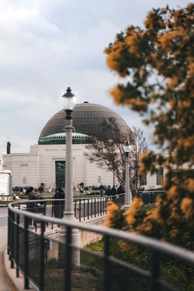 A large observatory building with a dome roof is seen in the background, surrounded by trees. The foreground features a railing and a lamp post, with some individuals walking and gathering outside the observatory.