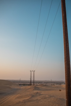 Power lines stretch across a vast desert landscape with sandy dunes in the foreground. A person walks along the sandy path, and in the distance, small structures and greenery are visible under the pale blue sky.