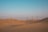 A vast desert landscape with sand dunes under a clear blue sky. A row of utility poles with power lines stretches across the sandy terrain. A small group of camels is visible in the distance, walking along the dunes.