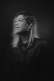 A black and white portrait of a woman with shoulder-length hair looking upwards. The lighting creates a soft, ethereal effect, with part of her face in focus while the other part is blurred, giving a sense of motion or dream-like quality.