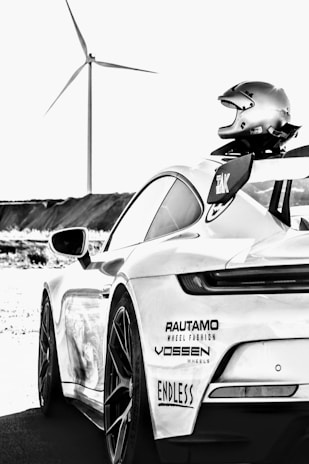 A sleek sports car is shown from the rear side with a racing helmet placed on its spoiler. The car has several stickers from brands such as Rautamo, Vossen, and Endless. In the background, there is a wind turbine set against a clear sky, indicating an open, possibly rural setting.