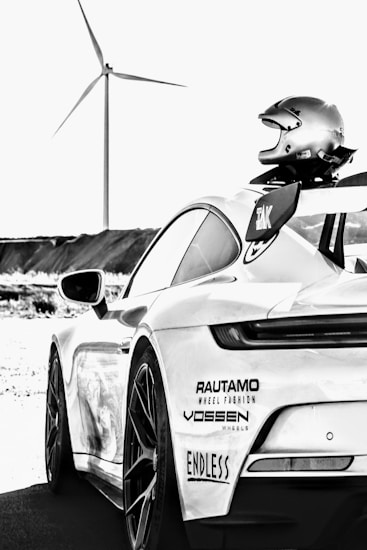 A sleek sports car is shown from the rear side with a racing helmet placed on its spoiler. The car has several stickers from brands such as Rautamo, Vossen, and Endless. In the background, there is a wind turbine set against a clear sky, indicating an open, possibly rural setting.