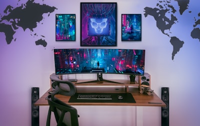 A modern gaming setup features a large curved monitor displaying a futuristic cyberpunk cityscape. The wooden desk includes a sleek black chair, stereo speakers, and a small model car. Above the monitor, three framed art pieces with cyberpunk aesthetics adorn the wall. The wall is also decorated with silhouetted world maps on either side.