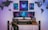 A modern gaming setup features a large curved monitor displaying a futuristic cyberpunk cityscape. The wooden desk includes a sleek black chair, stereo speakers, and a small model car. Above the monitor, three framed art pieces with cyberpunk aesthetics adorn the wall. The wall is also decorated with silhouetted world maps on either side.