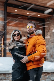 A couple stands outdoors in a snowy setting, both wearing stylish winter jackets. The man, in an orange puffer jacket with a fur-lined hood, has sunglasses with reflective lenses and a beard. The woman, in a black jacket, also wears sunglasses and has long hair. They are embracing each other, creating a warm and intimate atmosphere against a backdrop of buildings with wooden and brick features.