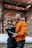 A couple stands outdoors in a snowy setting, both wearing stylish winter jackets. The man, in an orange puffer jacket with a fur-lined hood, has sunglasses with reflective lenses and a beard. The woman, in a black jacket, also wears sunglasses and has long hair. They are embracing each other, creating a warm and intimate atmosphere against a backdrop of buildings with wooden and brick features.