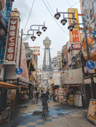 A bustling urban street scene in Japan with vibrant signs and advertisements in Japanese. A distinct tower rises in the background, while pedestrians and cyclists move along the street. The area is filled with shops, restaurants, and various decorative elements, creating a lively atmosphere.