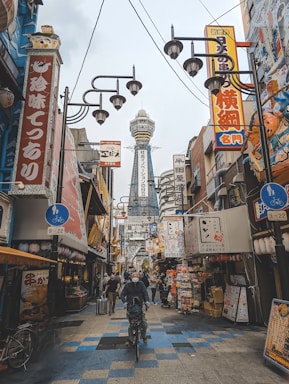 A bustling urban street scene in Japan with vibrant signs and advertisements in Japanese. A distinct tower rises in the background, while pedestrians and cyclists move along the street. The area is filled with shops, restaurants, and various decorative elements, creating a lively atmosphere.