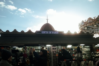 A market stall with a sign reading 'Gifts of Paris' is captured against a bright sky. The stall is filled with various souvenirs, including sunglasses and trinkets. Several people are browsing or taking photos, while a decorative carousel is partially visible on the right.