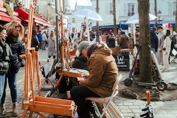 In a lively outdoor setting, artists present their work on easels while people browse and observe. A man sits on a chair, focused on his drawing or painting task. The scene is bustling with pedestrians walking along the cobblestone path, surrounded by buildings decorated with colorful red awnings and winter holiday decorations.
