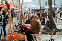In a lively outdoor setting, artists present their work on easels while people browse and observe. A man sits on a chair, focused on his drawing or painting task. The scene is bustling with pedestrians walking along the cobblestone path, surrounded by buildings decorated with colorful red awnings and winter holiday decorations.