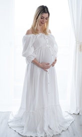 a pregnant woman in a white dress standing in front of a window