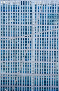 A modern building facade featuring a grid pattern of blue-tinted windows and white frames, intersected by three diagonal lines that add a dynamic visual element.