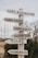A wooden signpost outdoors features several directional signs, each pointing to different locations such as a loft restaurant, farm store, chef's garden, dairy bar, tours, and milking parlor. The signpost is surrounded by natural elements like bushes and tall grass with a muted, overcast sky in the background.