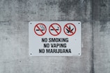 A rectangular sign on a concrete wall displays prohibitory symbols. Three red circles with diagonal slashes indicate no smoking, no vaping, and no marijuana. The words 'NO SMOKING,' 'NO VAPING,' and 'NO MARIJUANA' are boldly printed below the symbols.