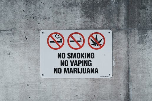 A rectangular sign on a concrete wall displays prohibitory symbols. Three red circles with diagonal slashes indicate no smoking, no vaping, and no marijuana. The words 'NO SMOKING,' 'NO VAPING,' and 'NO MARIJUANA' are boldly printed below the symbols.