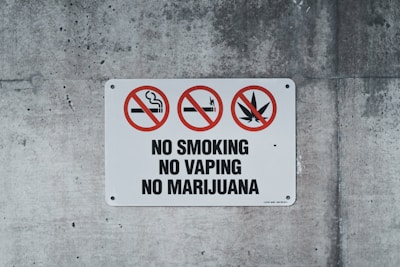 A rectangular sign on a concrete wall displays prohibitory symbols. Three red circles with diagonal slashes indicate no smoking, no vaping, and no marijuana. The words 'NO SMOKING,' 'NO VAPING,' and 'NO MARIJUANA' are boldly printed below the symbols.