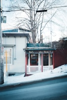 A small storefront with a rustic charm, featuring a red door and white trim. The sign above the entrance reads 'Herbs Garden Thing.' The building is nestled next to a snowy sidewalk on a sloping street, with bare trees in the background.