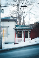 A small storefront with a rustic charm, featuring a red door and white trim. The sign above the entrance reads 'Herbs Garden Thing.' The building is nestled next to a snowy sidewalk on a sloping street, with bare trees in the background.