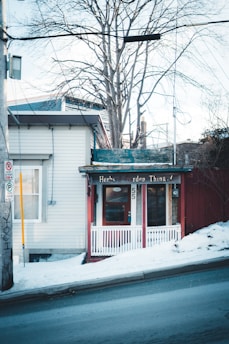 A small storefront with a rustic charm, featuring a red door and white trim. The sign above the entrance reads 'Herbs Garden Thing.' The building is nestled next to a snowy sidewalk on a sloping street, with bare trees in the background.
